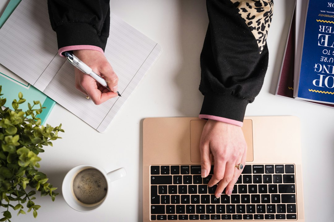 A person's hands writing notes in a journal beside an open laptop and a cup of coffee