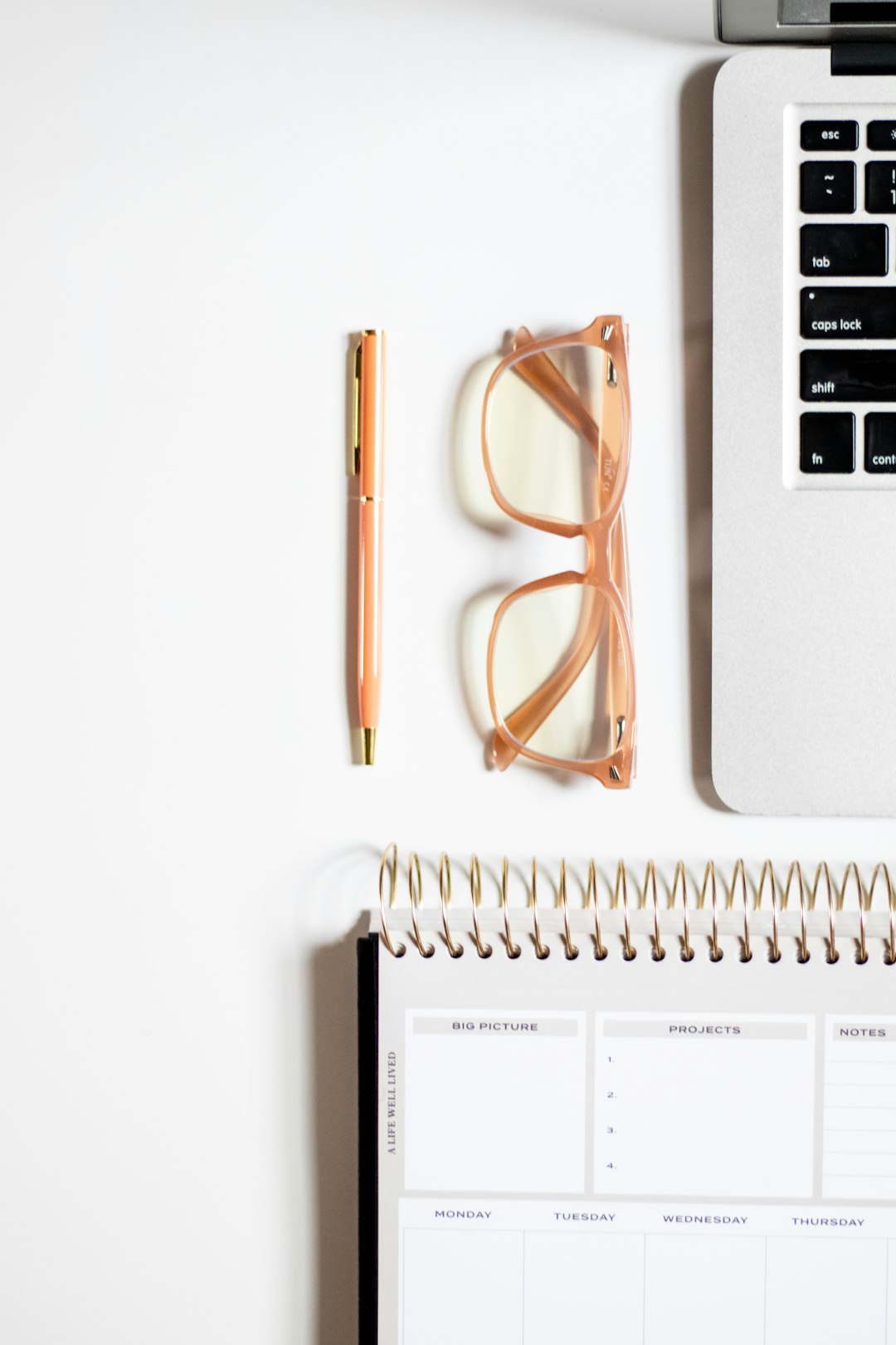 A tidy flatlay of a laptop, reading glasses, pen, spiral notebook, and calendar on a white desk